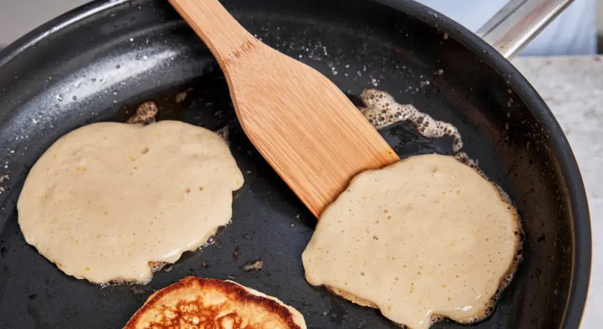 Buttermilch pancakes braten in der Pfanne. Ein Holzspatel wendet einen goldbraunen Pancake.