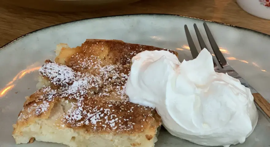 Ein Stück saftiger Apfelkuchen, bestäubt mit Puderzucker, serviert mit einer großen Portion Schlagsahne und einer Gabel.