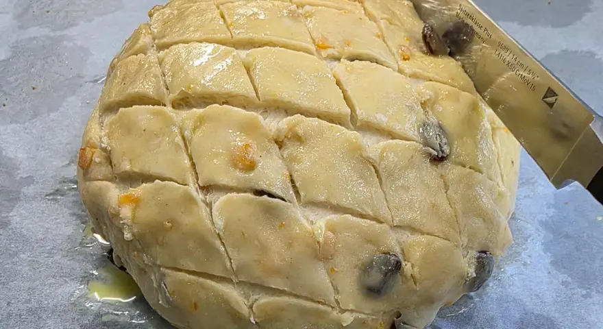 Ein runder Teigling mit eingeschnittenen Rauten, bereit für das Osterfladen Rezept. Rosinen und Orangenschale sind sichtbar.