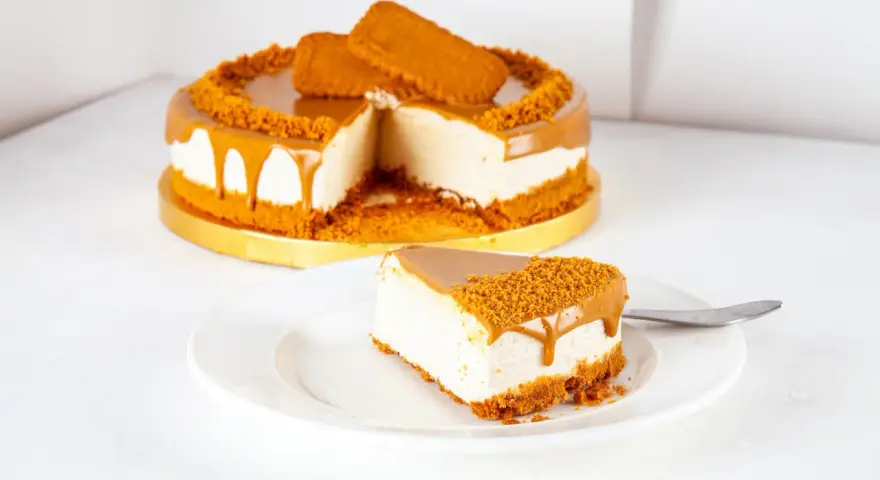 Ein cremiger Lotus Cheesecake mit Karamellglasuren und Keksbröseln. Ein Stück auf einem Teller, daneben ein ganzer Kuchen.