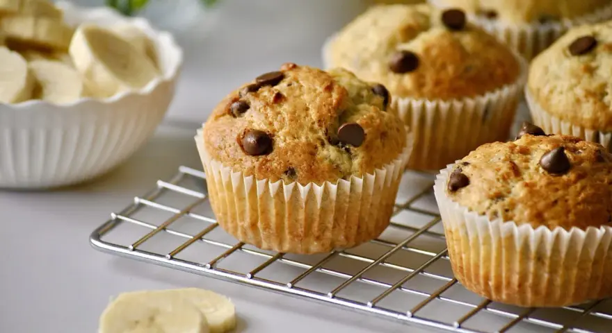 Frisch gebackene schoko bananen muffins mit Schokostückchen auf einem Gitter. Daneben eine Schüssel mit Bananenscheiben.