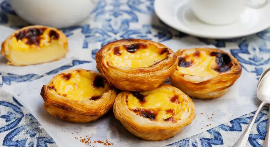 Vier köstliche Pastel de Nata auf einem blauen Tischtuch, mit einer Tasse Kaffee im Hintergrund.
