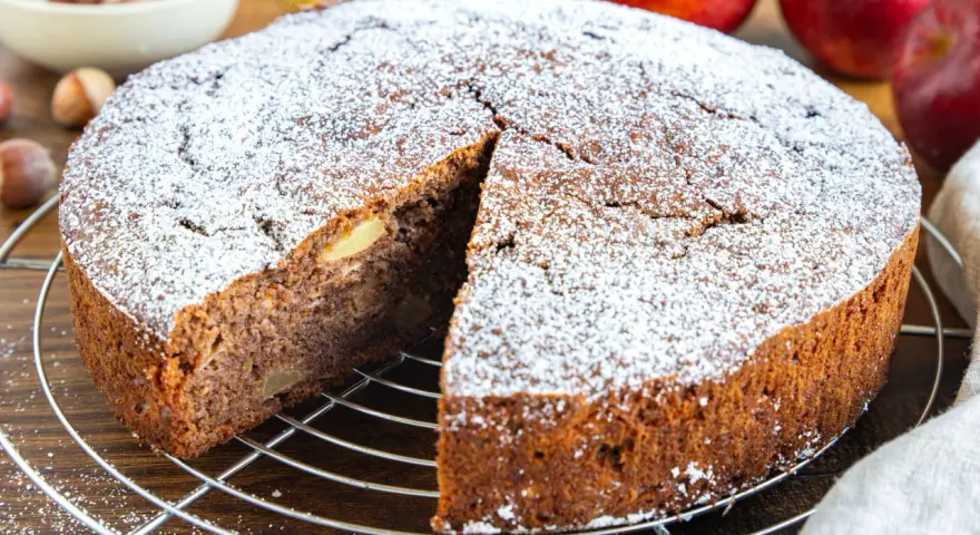 Ein saftiger russischer Apfelkuchen, bestäubt mit Puderzucker, wartet auf seine Genießer. Apfelstücke sind im dunklen Teig sichtbar.