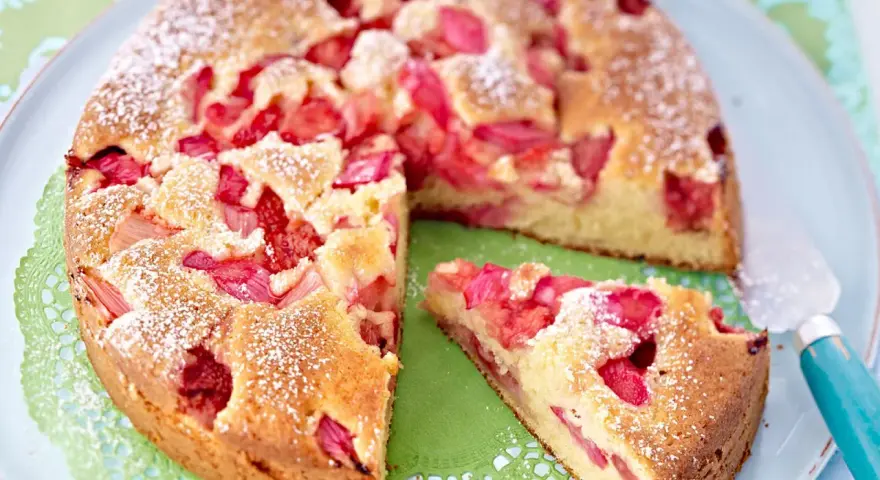 Ein saftiger Erdbeer Rhabarber Kuchen, bestäubt mit Puderzucker, liegt auf einem Teller. Ein Stück wurde bereits entnommen.