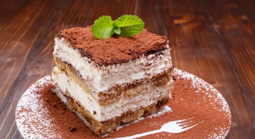 Ein Stück cremiges Tiramisu ohne Ei, bestäubt mit Kakao und garniert mit Minze, auf einem Teller mit Kakaopulver und einer Gabel.
