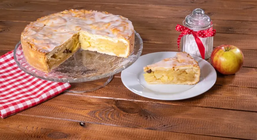 Omas weltbester Apfelkuchen, saftig und mit Zuckerguss überzogen, wartet auf einem Tortenständer. Ein Stück auf einem Teller und ein Apfel daneben laden zum Genießen ein.