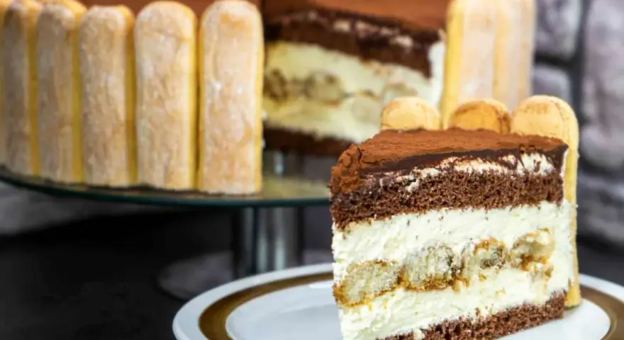 Ein Stück Tiramisu Torte mit Löffelbiskuits und Kakao bestäubt. Im Hintergrund die ganze Torte, ebenfalls mit Löffelbiskuits umrandet.
