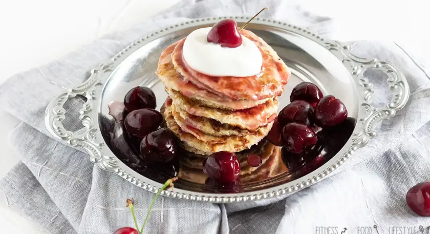 Ein Stapel Bananen Pancakes mit Kirschen und Sahne auf einem Silbertablett, garniert mit frischen Kirschen.