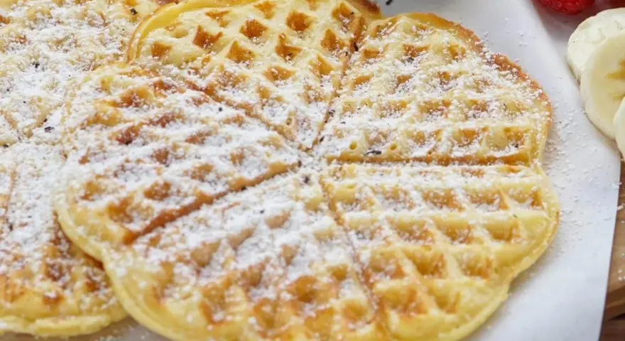 Zuckerbestäubte Buttermilchwaffeln, die auf einem Holzbrett liegen. Daneben sind Himbeeren und Bananenscheiben zu sehen.