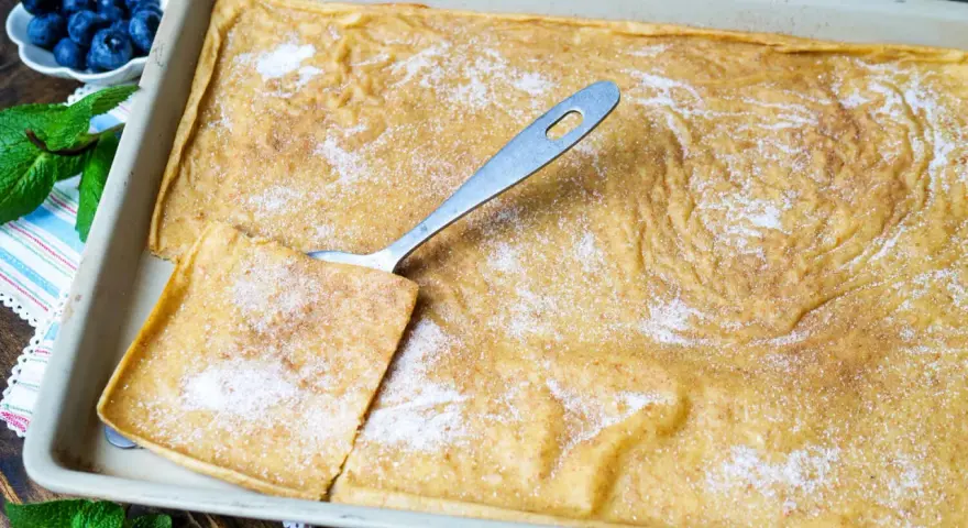 Ein frisch gebackener Ofenpfannkuchen, bestreut mit Zucker und Zimt, bereit zum Servieren. Ein Stück wird mit einem Löffel herausgehoben.
