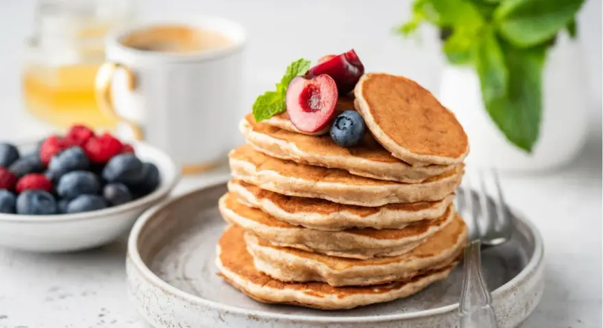 Ein Stapel veganer Pancakes, garniert mit Kirschen und Blaubeeren, daneben eine Schale Beeren und eine Tasse Kaffee.