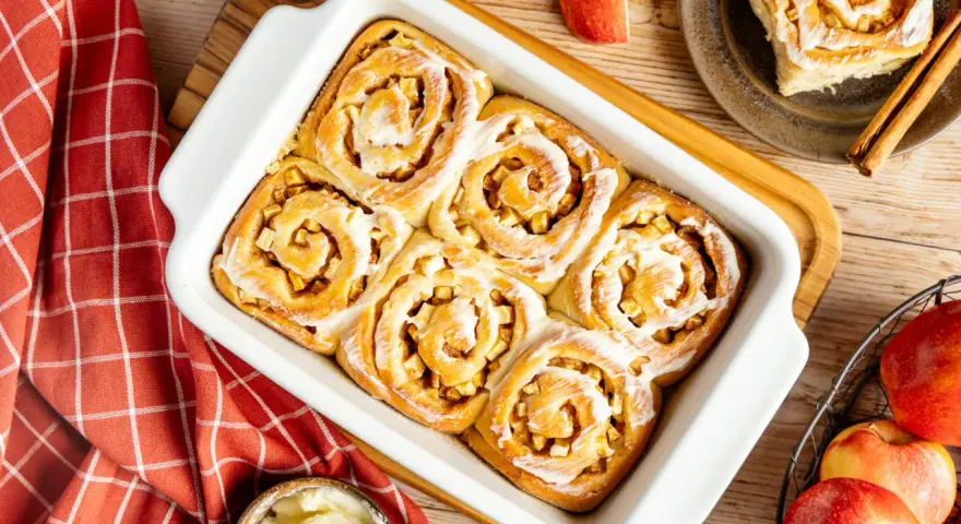 Frisch gebackene Apfel Zimt Schnecken mit Zuckerguss in einer Auflaufform. Daneben Äpfel und Zimtstangen.