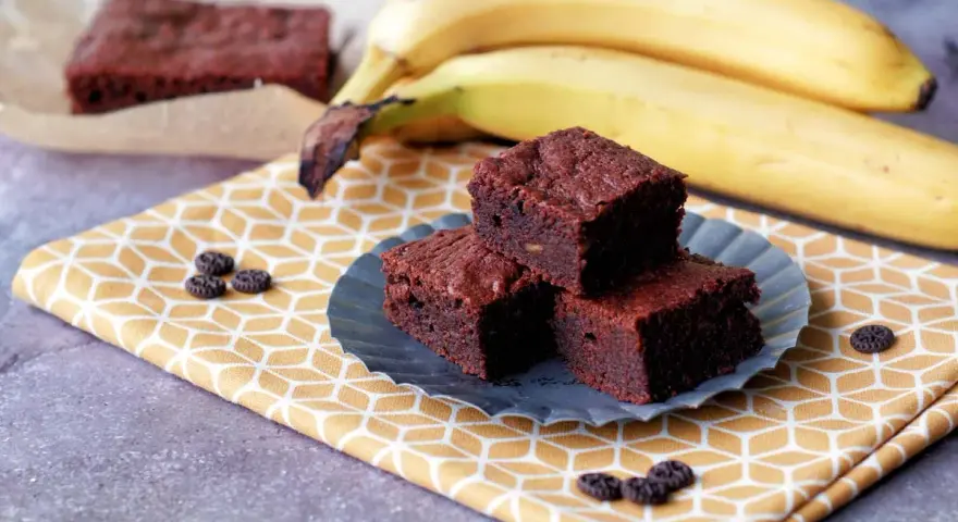 Saftige Bananen Brownies auf einem gelben Tuch, daneben reife Bananen. Ein Genuss!