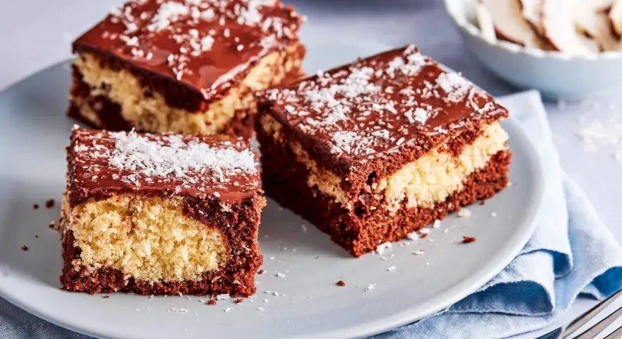 Drei Stücke saftiger Schoko Kokos Kuchen mit Schokoladenglasur und Kokosraspeln auf einem grauen Teller.