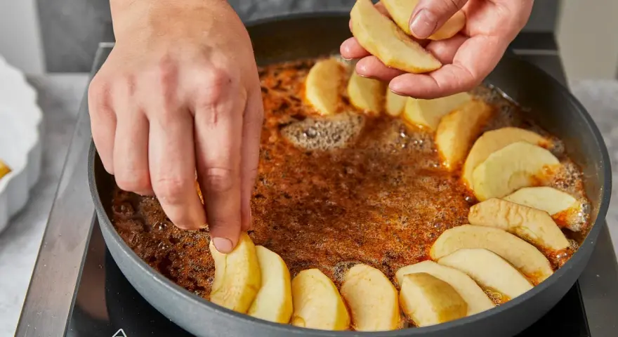Hände legen Apfelspalten in eine Pfanne mit karamellisierter Butter. Die Zubereitung der klassischen Tarte Tatin beginnt.