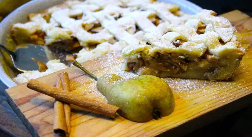 Ein köstlicher Birnenkuchen mit Gittermuster, bestäubt mit Puderzucker. Daneben liegen eine Birne und Zimtstangen – perfekt für ein Birnenkuchen Rezept.