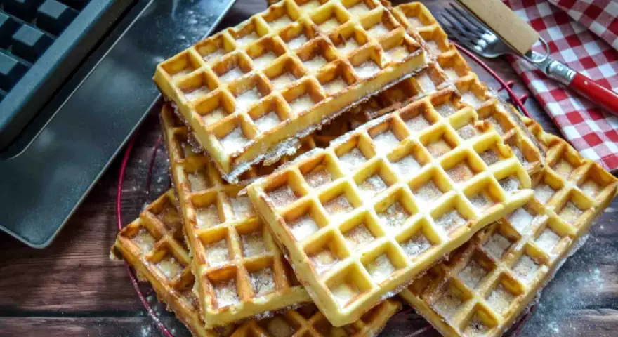 Frisch gebackene belgische Waffeln, mit Puderzucker bestäubt, liegen auf einem Gitter. Daneben ein Waffeleisen und eine karierte Serviette.