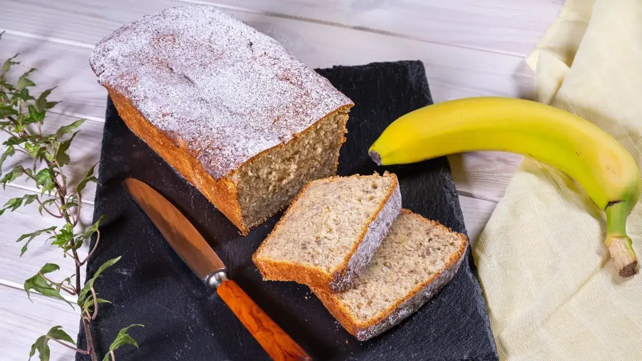 Ein saftiger Bananenkuchen, best&auml;ubt mit Puderzucker, liegt auf einem schwarzen Stein. Daneben ein Messer und eine Banane &ndash; perfekt f&uuml;r ein schnelles Bananenkuchen Rezept.