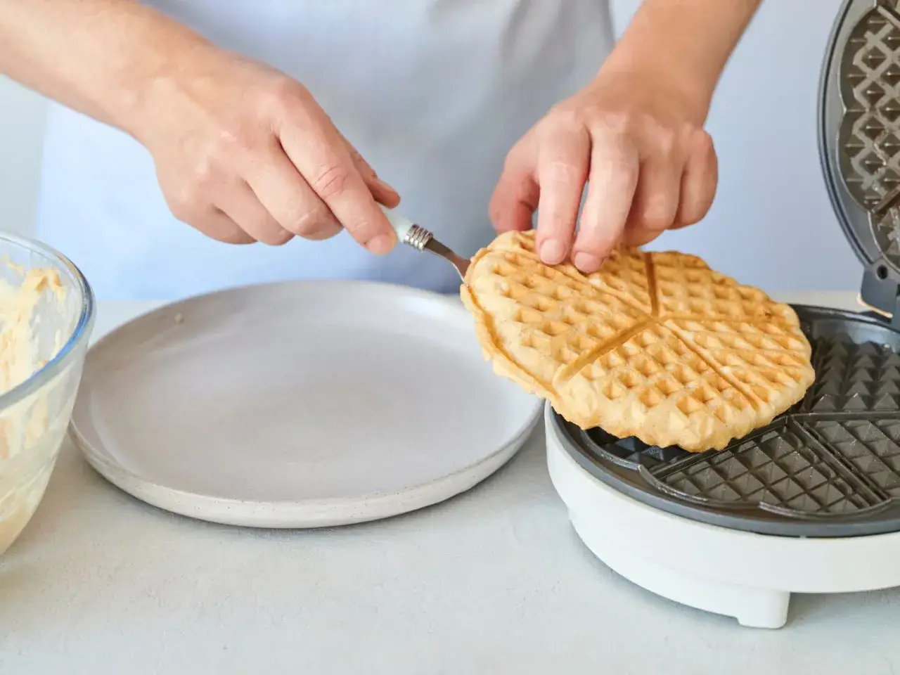 Hände nehmen eine frisch gebackene Dinkel Waffel aus dem Waffeleisen. Daneben liegen Teig und ein Teller.