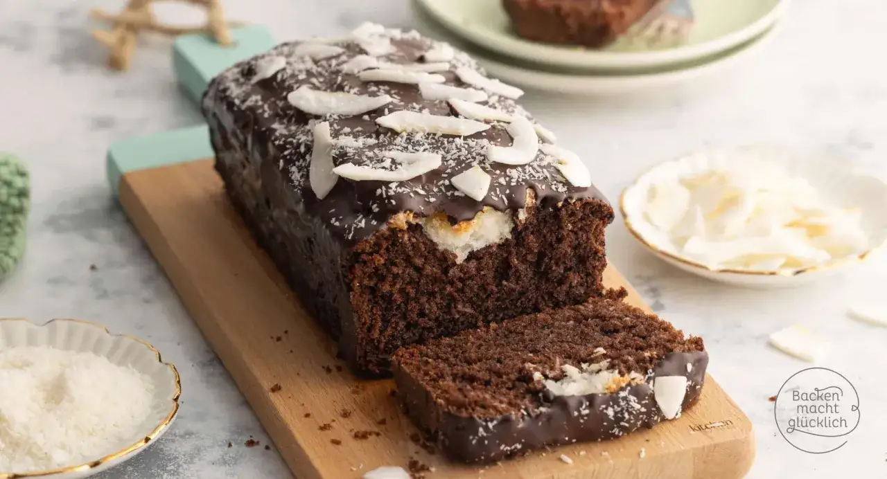 Schoko Kokos Kuchen mit dunkler Glasur und Kokosraspeln, ein Stück angeschnitten, auf einem Holzbrett.
