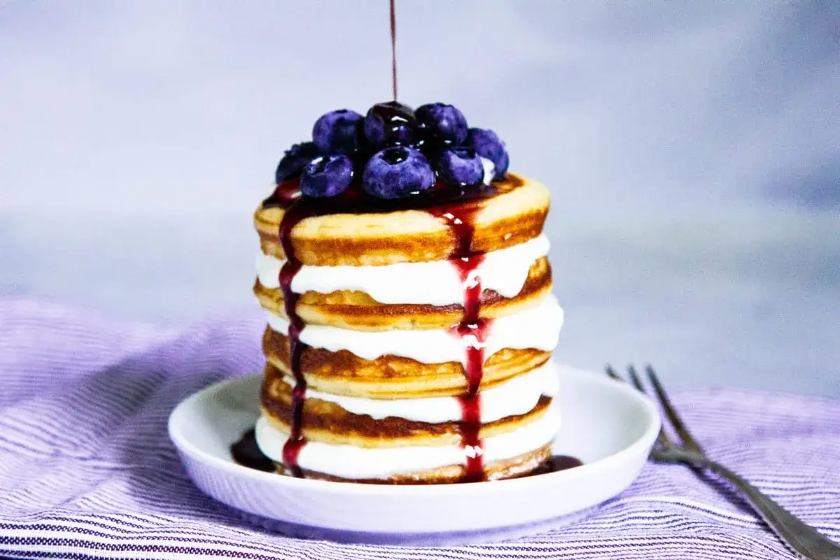Ein Stapel glutenfreier Pfannkuchen mit Sahne, Blaubeeren und Sirup, serviert auf einem Teller mit einer Gabel.