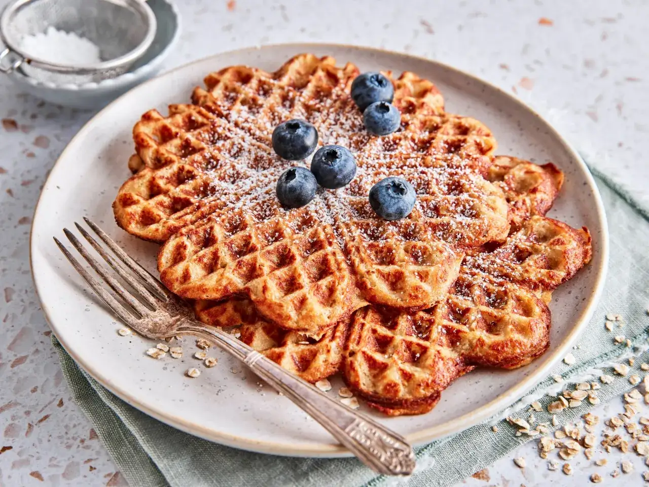 Goldbraune Haferflocken Waffeln, best&auml;ubt mit Puderzucker und garniert mit frischen Blaubeeren, auf einem Teller mit einer Gabel.