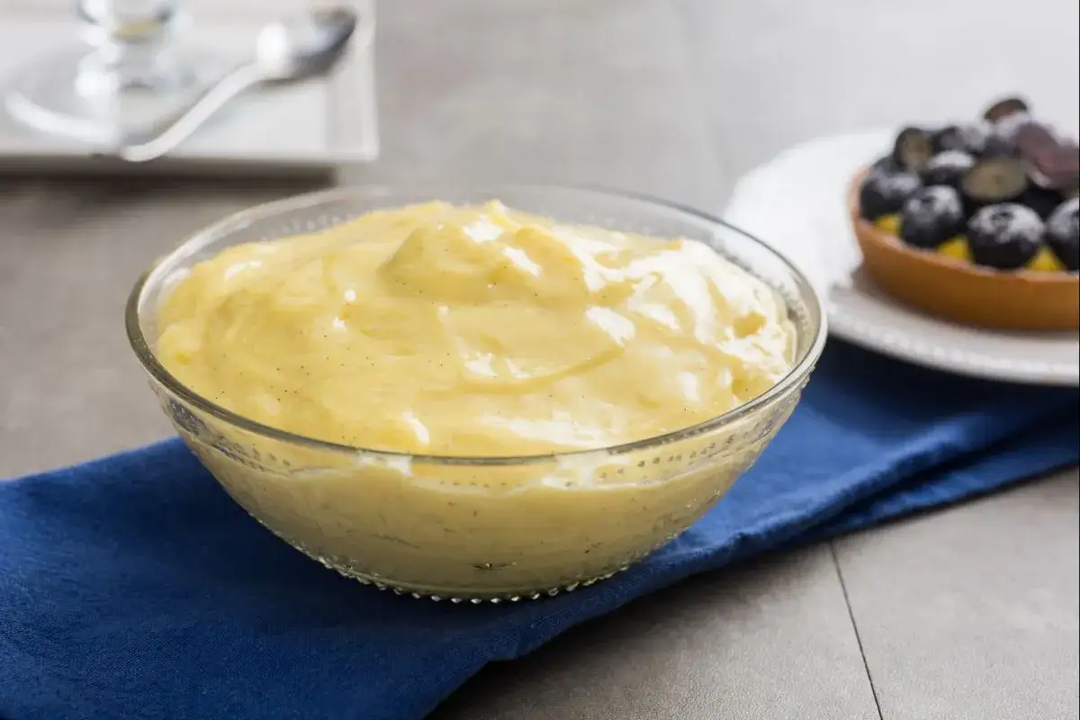 Cremige Crema Pasticcera in einer Glasschüssel, bereit zum Genießen. Daneben ein kleiner Kuchen mit Blaubeeren.