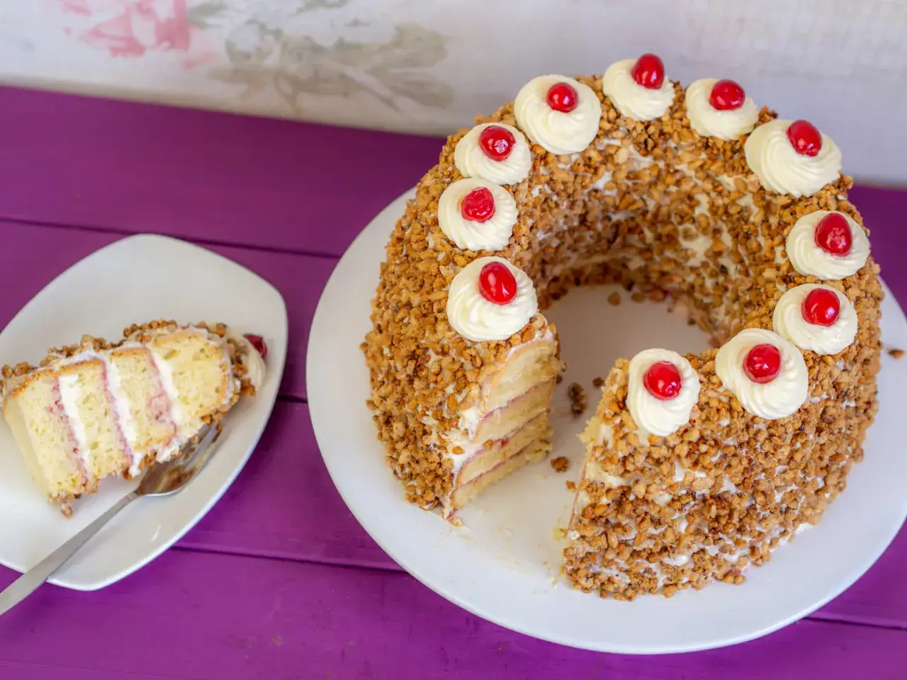 Ein köstlicher Frankfurter Kranz, umhüllt von Nüssen und verziert mit Sahnetuffs und Kirschen. Ein Stück ist bereits angeschnitten.
