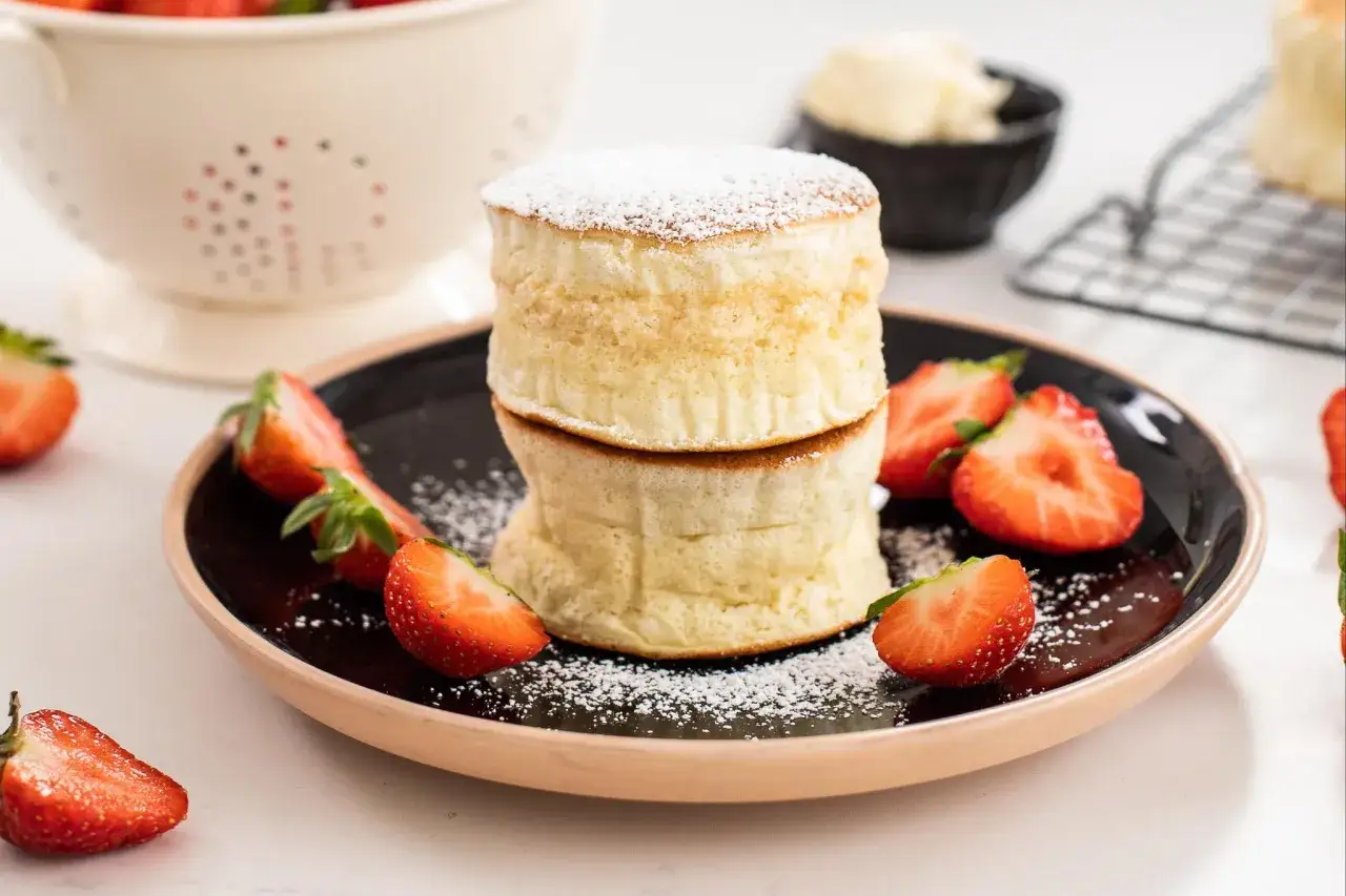 Zwei fluffige japanische Pancakes, bestäubt mit Puderzucker, serviert mit frischen Erdbeeren auf einem schwarzen Teller.