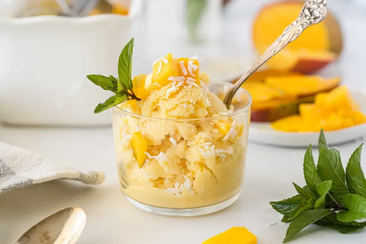 Cremiges Mango Eis mit Kokosraspeln und frischer Minze, garniert mit Mangostücken. Ein Löffel wartet schon auf den Genuss.