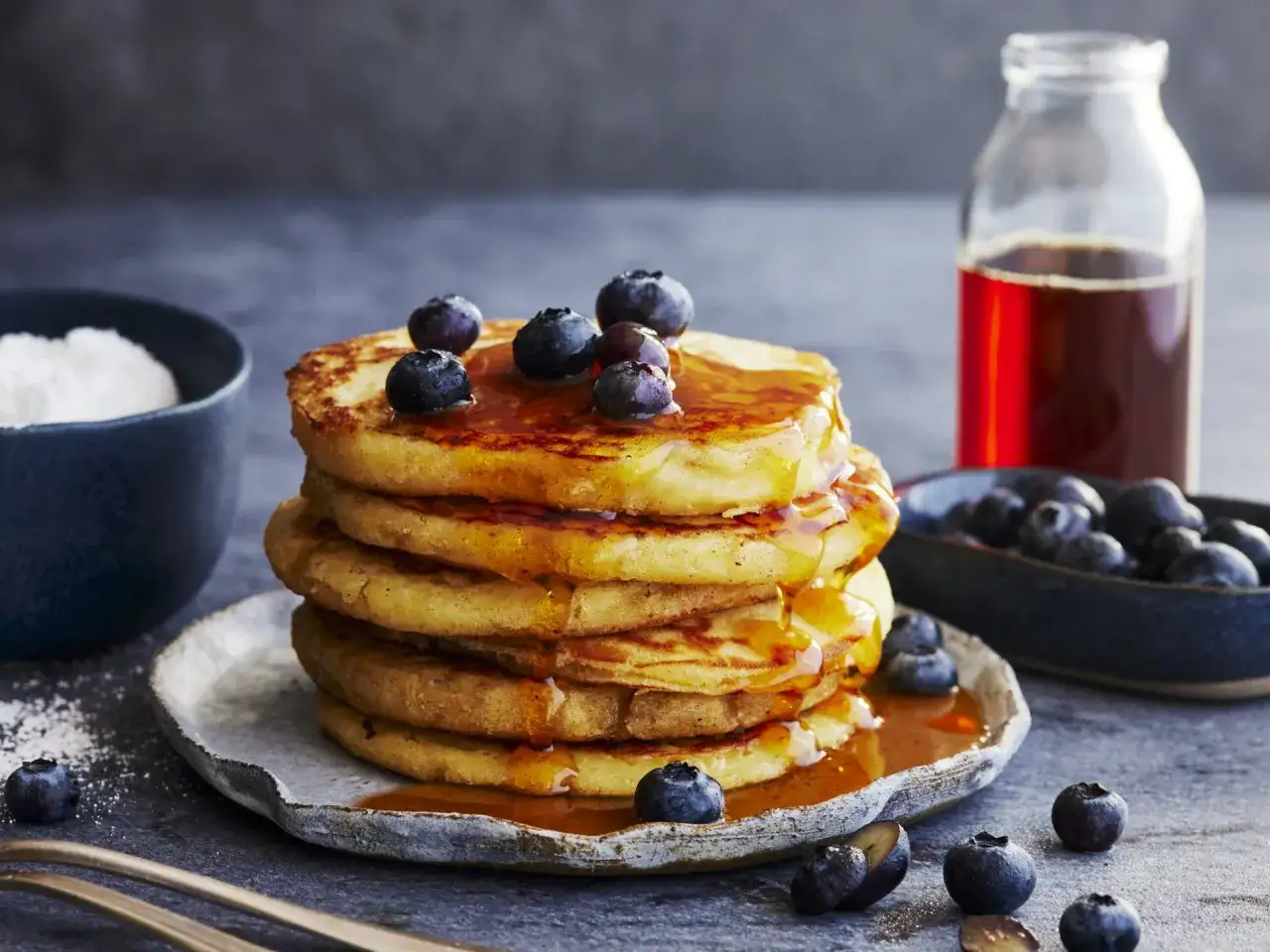 Ein Stapel Buttermilch-Pancakes, beträufelt mit Sirup und garniert mit Blaubeeren. Daneben eine Flasche Sirup und eine Schüssel mit Blaubeeren.