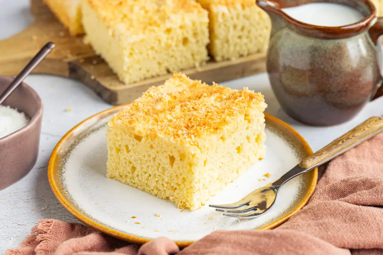 Ein Stück saftiger Buttermilch Kuchen mit Kokosraspeln auf einem Teller, daneben eine Kanne Milch und eine Schüssel mit Kokosraspeln.
