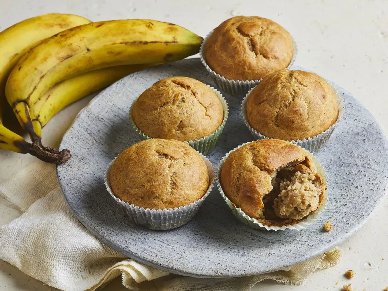Saftige bananen muffins auf einem grauen Teller, daneben reife Bananen. Einer der Muffins ist angebrochen und zeigt die feine Krume.