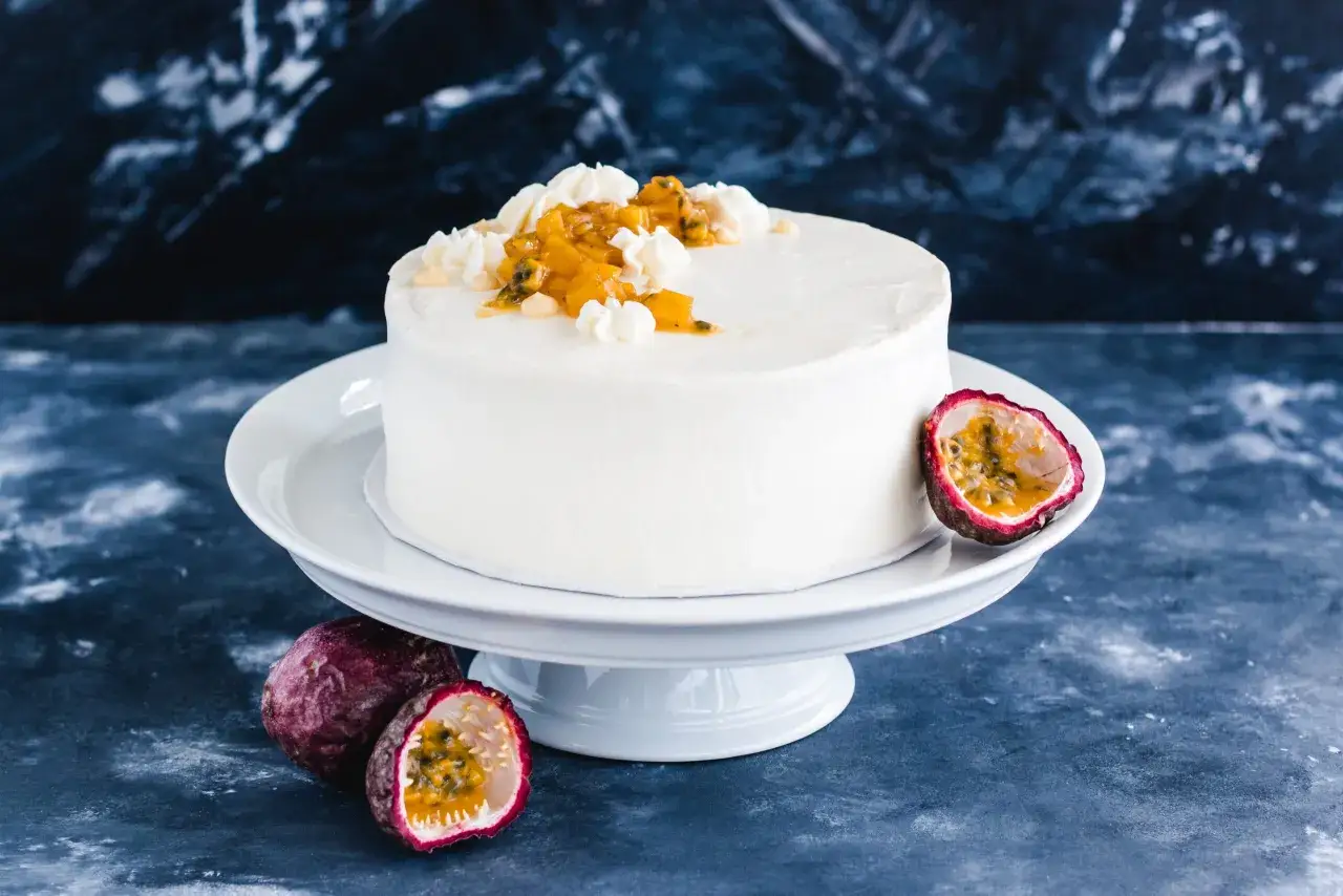 Eine weiße Maracuja Torte mit frischer Maracuja-Frucht und Sahnetupfern verziert, daneben liegen ganze und halbierte Maracujas.