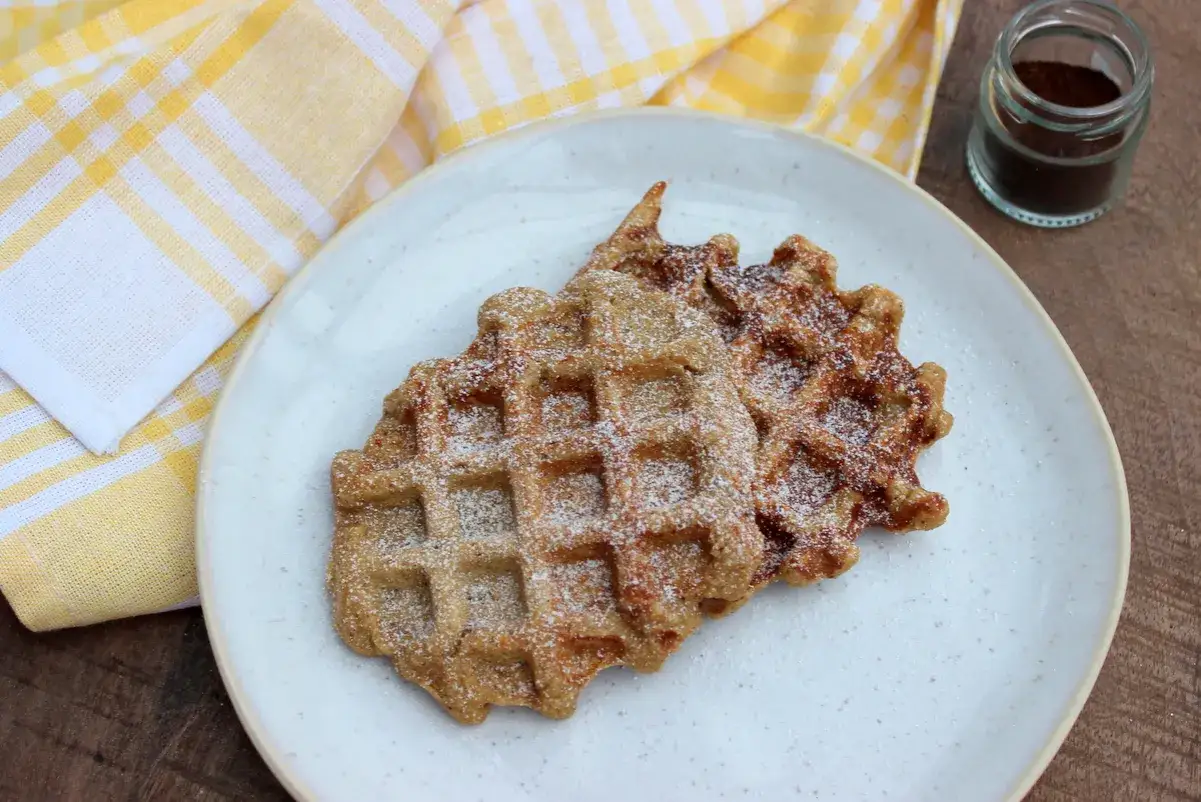 Zwei goldbraune Joghurt Waffeln, mit Puderzucker bestäubt, auf einem Teller. Daneben ein kleines Glas mit Kaffeepulver.