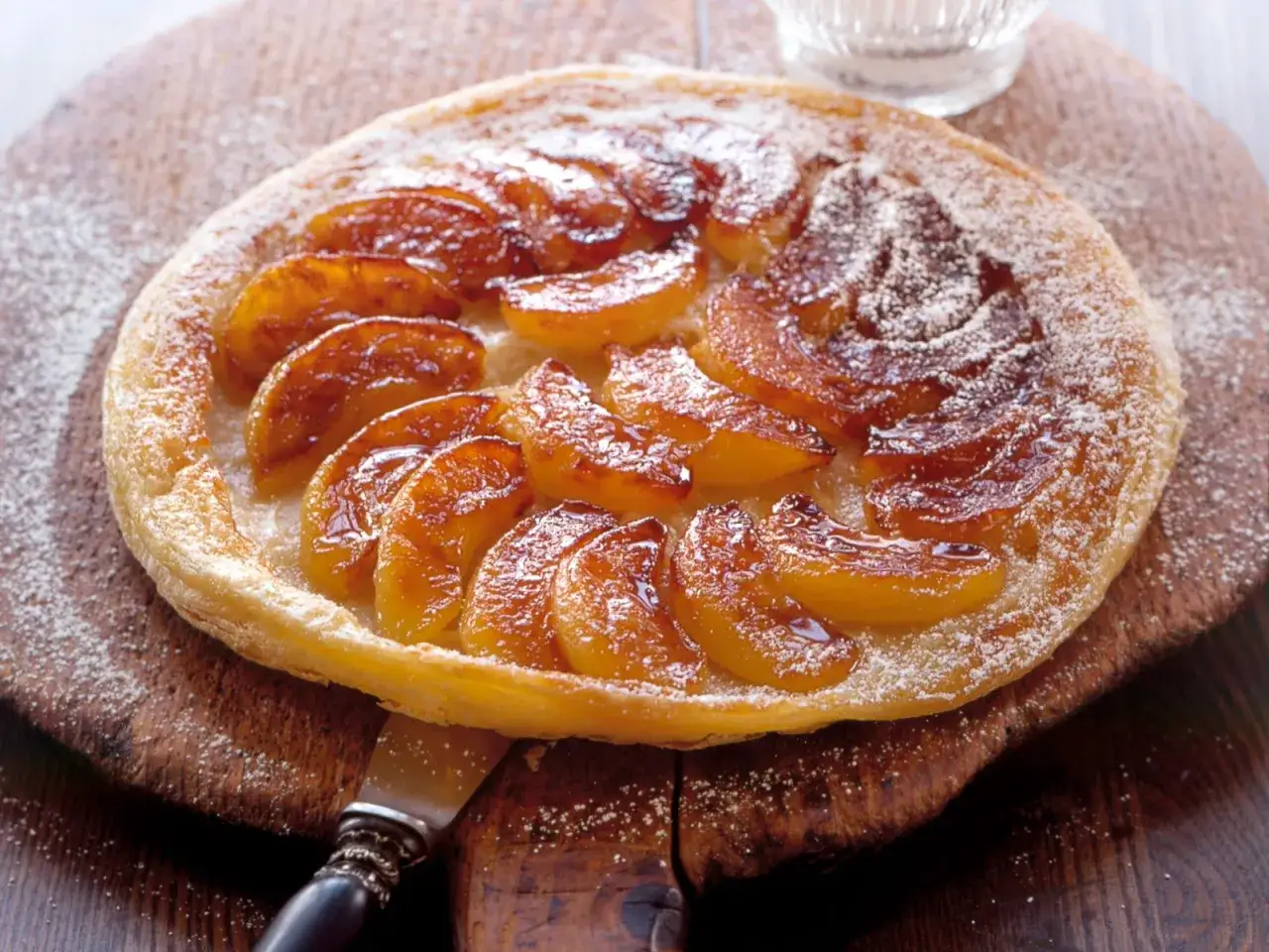 Eine karamellisierte Tarte Tatin mit saftigen Apfelspalten, bestäubt mit Puderzucker, auf einem rustikalen Holzbrett.