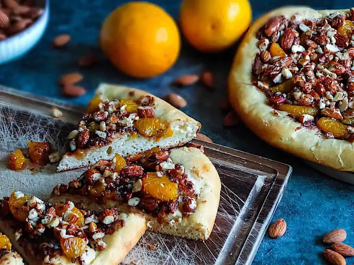 S&uuml;&szlig;e Nutella Pizza mit Mandeln und Orangenst&uuml;cken, auf einem Holzbrett serviert.