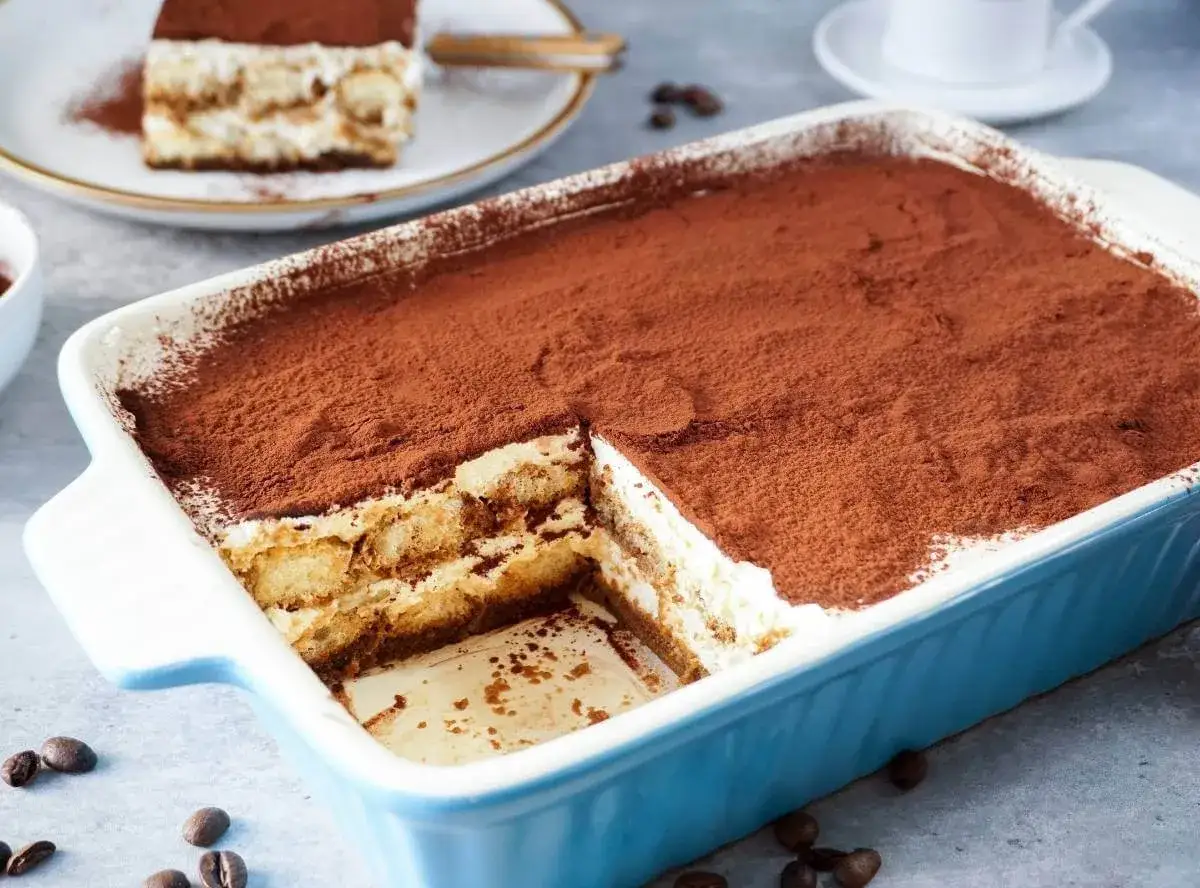 Ein köstliches Tiramisu ohne Ei, in einer blauen Auflaufform, bestäubt mit Kakao. Ein Stück ist bereits entnommen.