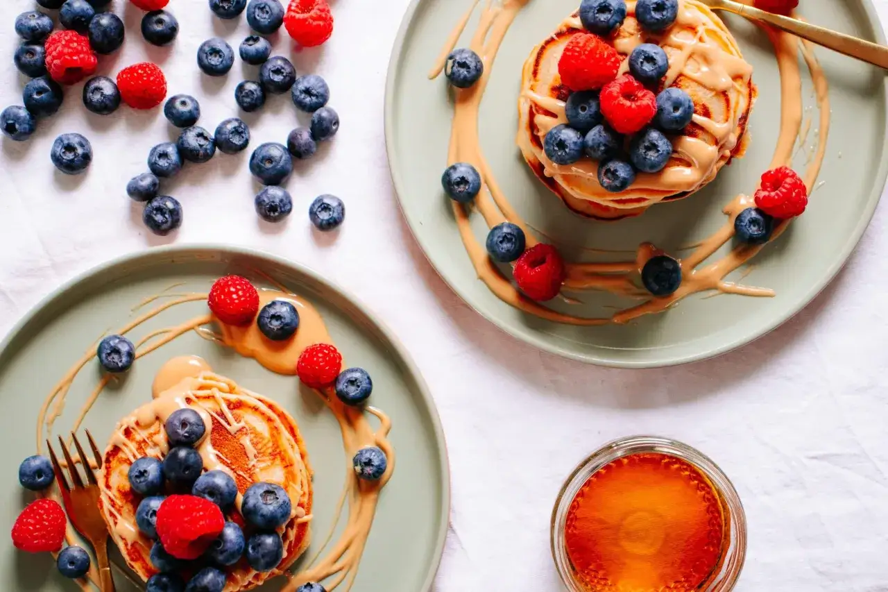 Zwei Teller mit leckeren, fluffigen bananen pancakes ohne ei, garniert mit Blaubeeren, Himbeeren und Erdnussbutter. Daneben ein Glas Honig.