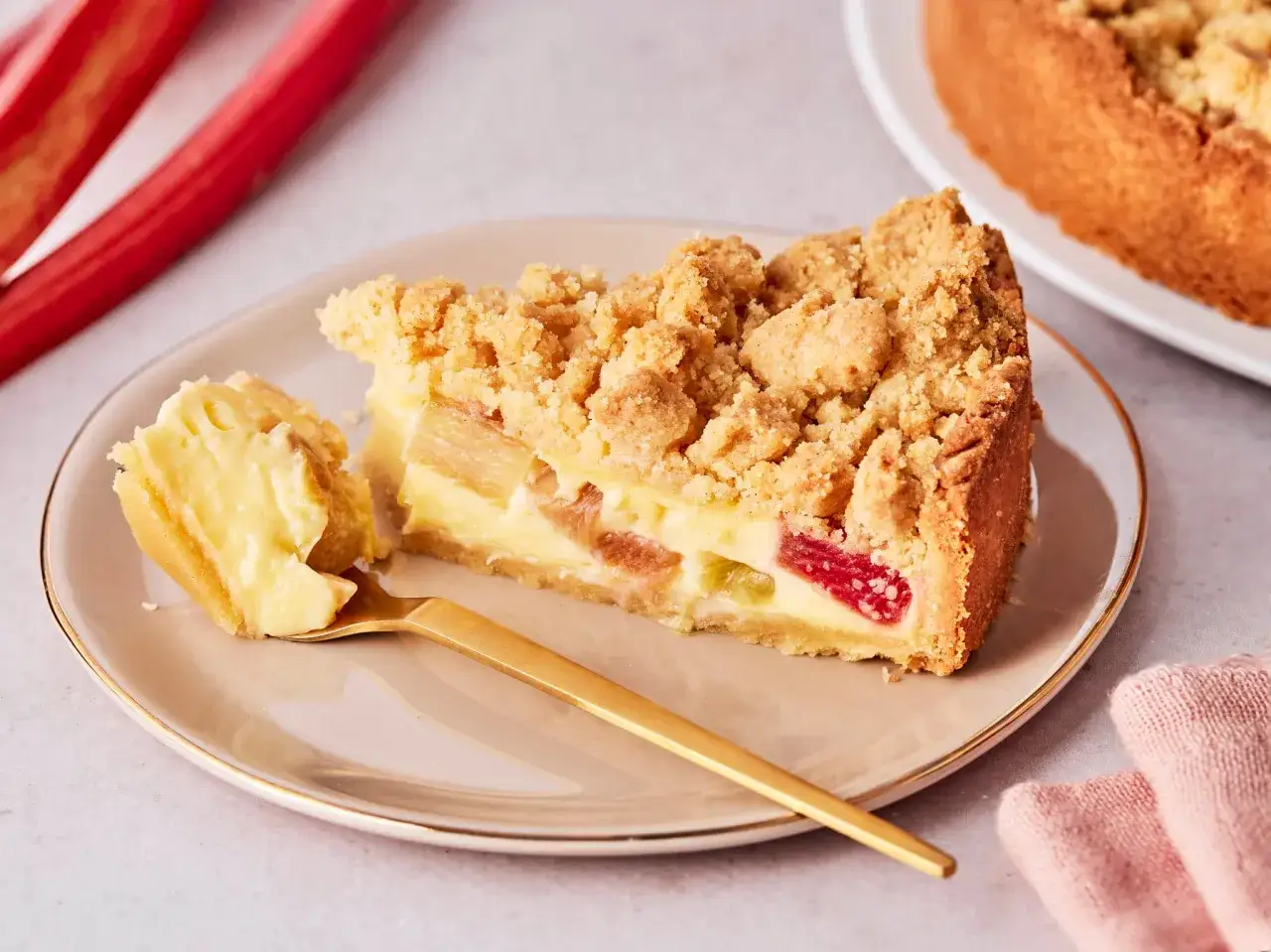 Ein Stück Rhabarberkuchen mit Pudding und Streuseln auf einem Teller, daneben ein goldener Löffel und rote Rhabarberstangen.