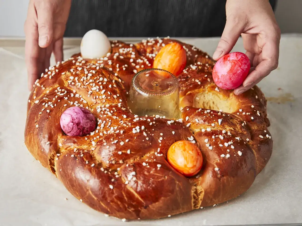 Hände schmücken einen goldbraunen Osterkranz mit bunten Eiern. Das Backen des Osterkranzes ist fast vollendet.