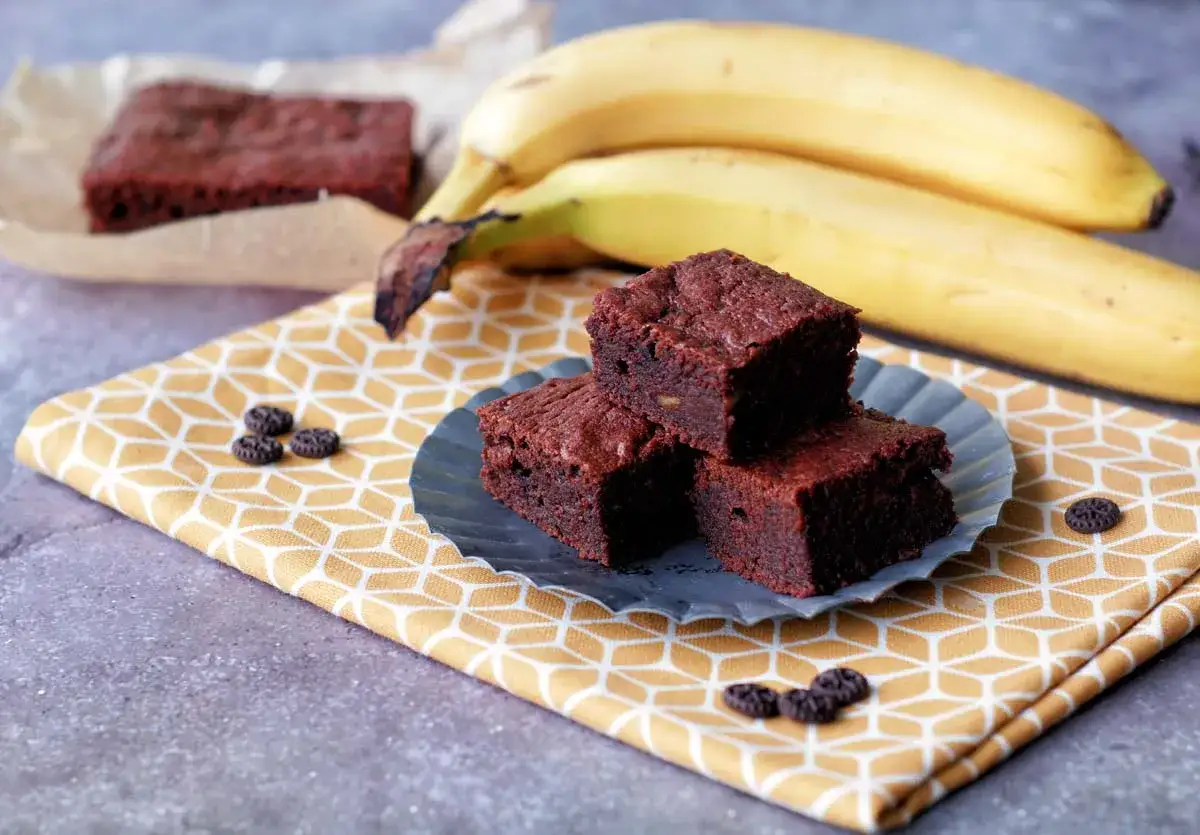 Saftige Bananen Brownies auf einem gelben Tuch, daneben reife Bananen. Ein Genuss!