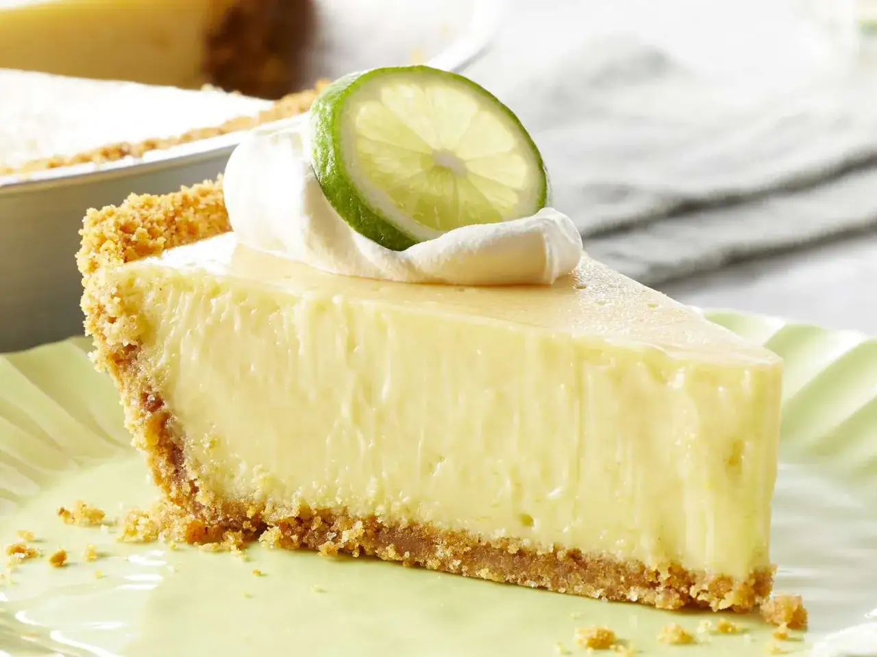 Ein cremiges St&uuml;ck Key Lime Pie mit Schlagsahne und Limettenscheibe auf einem hellgr&uuml;nen Teller.