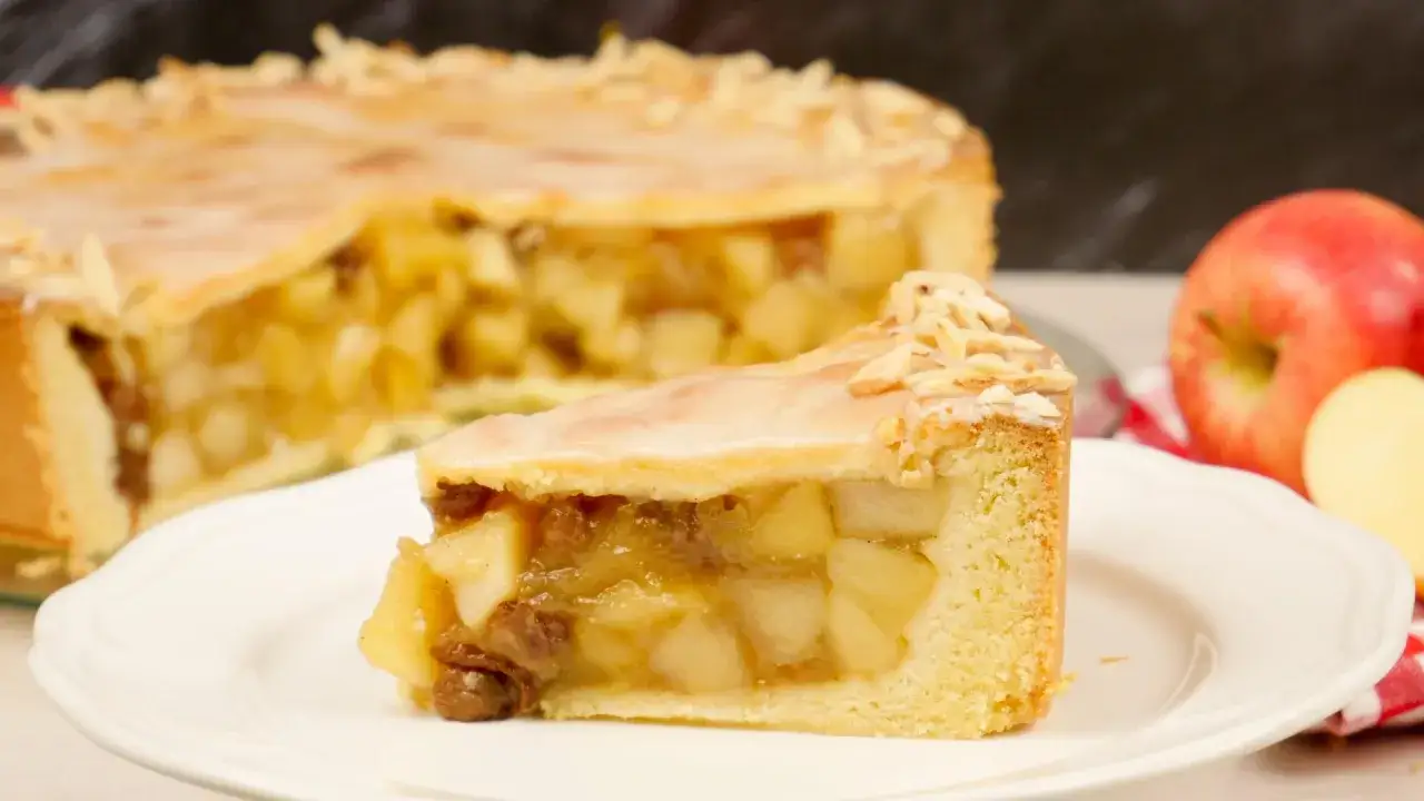 Ein St&uuml;ck gedeckter Apfelkuchen mit Mandelsplittern und Zuckerguss auf einem Teller, daneben ein ganzer Kuchen und &Auml;pfel.