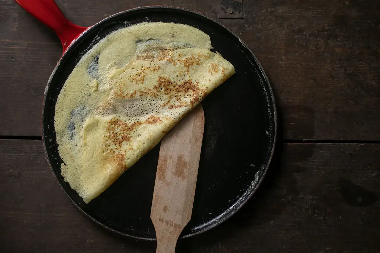 Ein goldbrauner Crêpe wird in einer Pfanne mit rotem Griff mit einem Holzspatel gewendet. Das perfekte Crêpes Rezept gelingt so.