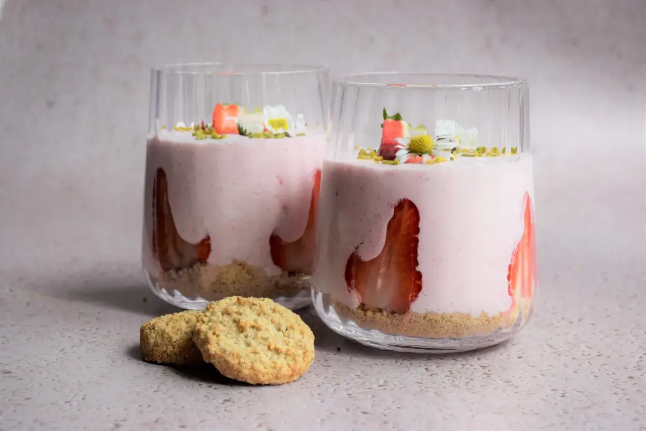 Zwei Gl&auml;ser mit cremigem Erdbeer-Dessert, garniert mit frischen Erdbeeren und G&auml;nsebl&uuml;mchen. Daneben liegen zwei knusprige Kekse.