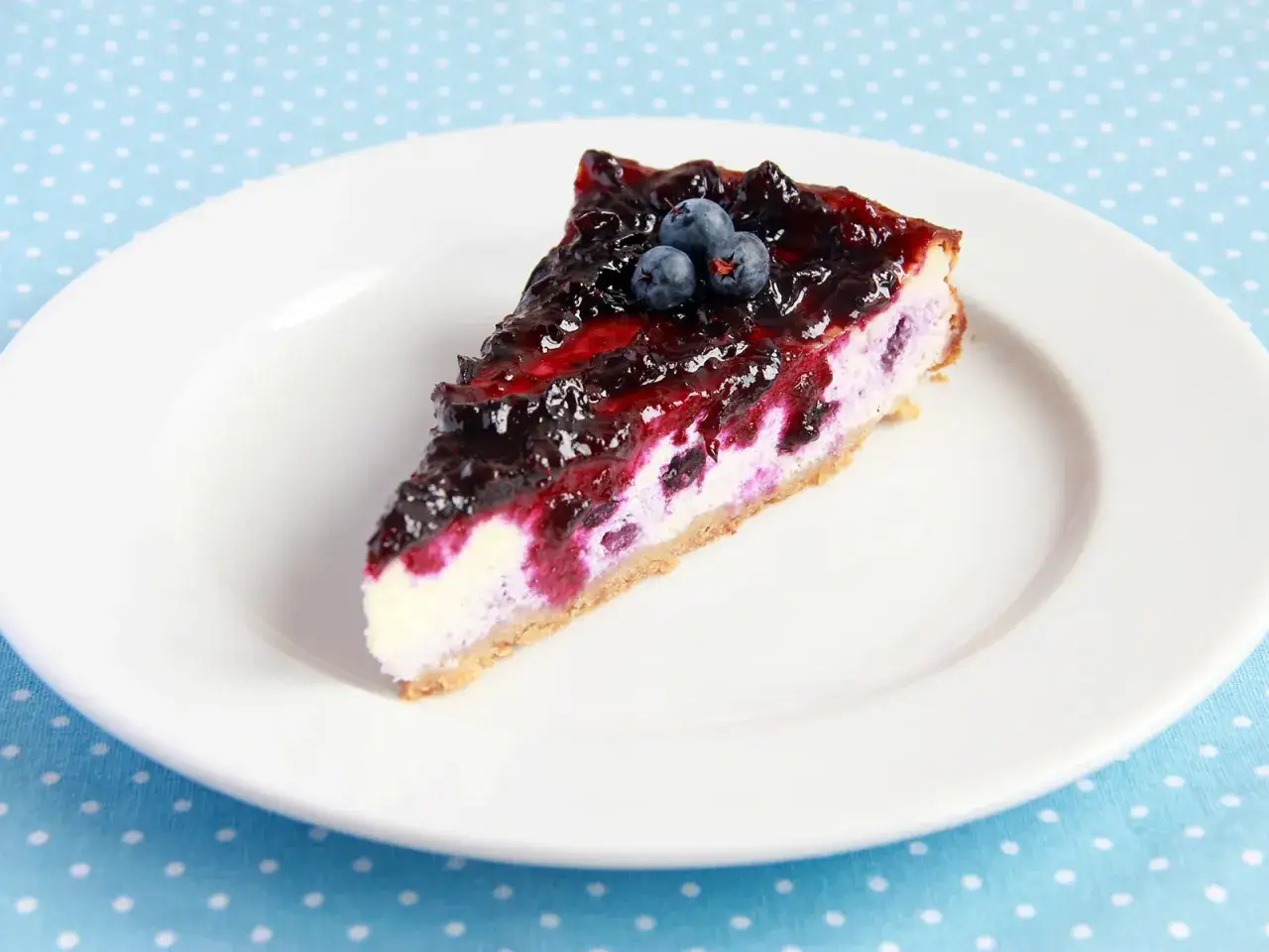 Ein St&uuml;ck cremiger Blueberry Cheesecake mit Blaubeersauce und frischen Beeren, serviert auf einem wei&szlig;en Teller.