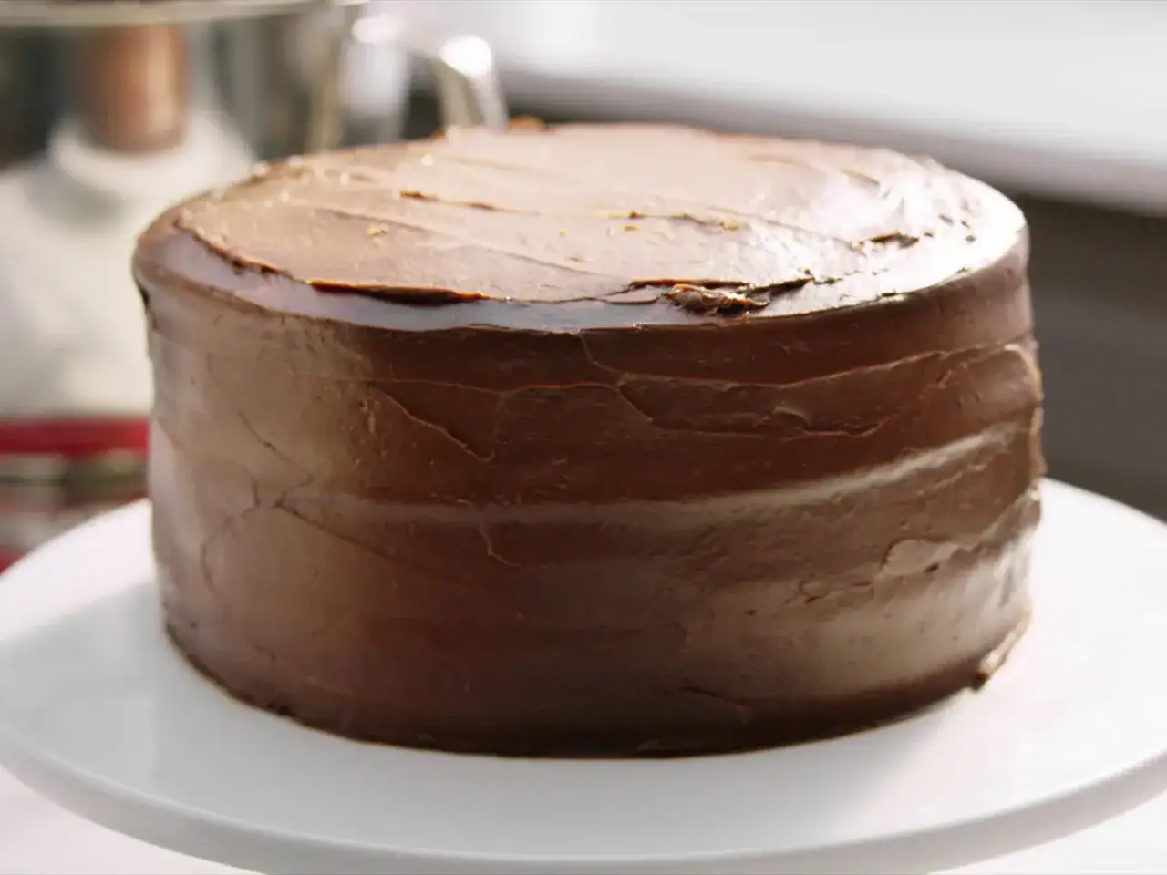 Eine köstliche, runde Schoko Torte mit dunkler Schokoladenglasur, die auf einem weißen Tortenständer steht.