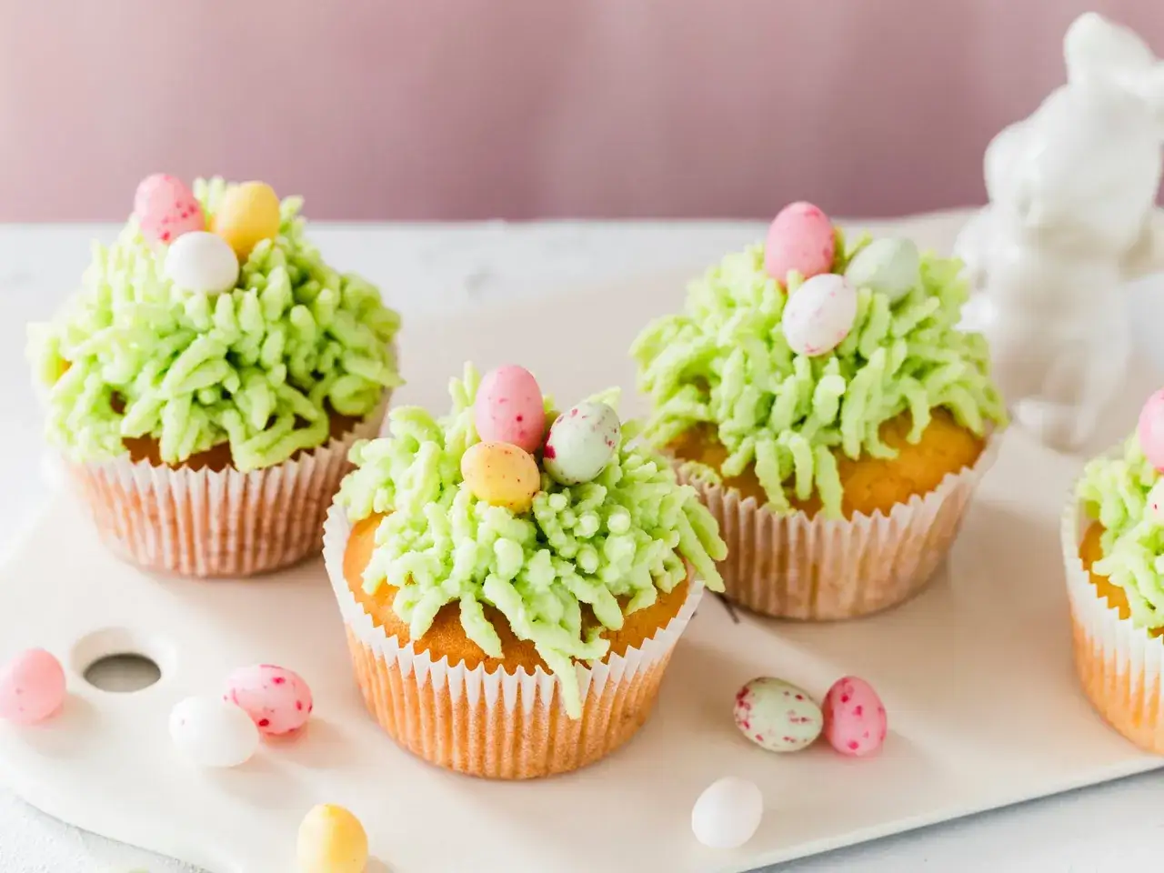 Oster Cupcakes mit grünem Zuckerguss, der wie Gras aussieht, und bunten Schokoladeneiern. Ein weißer Osterhase im Hintergrund.