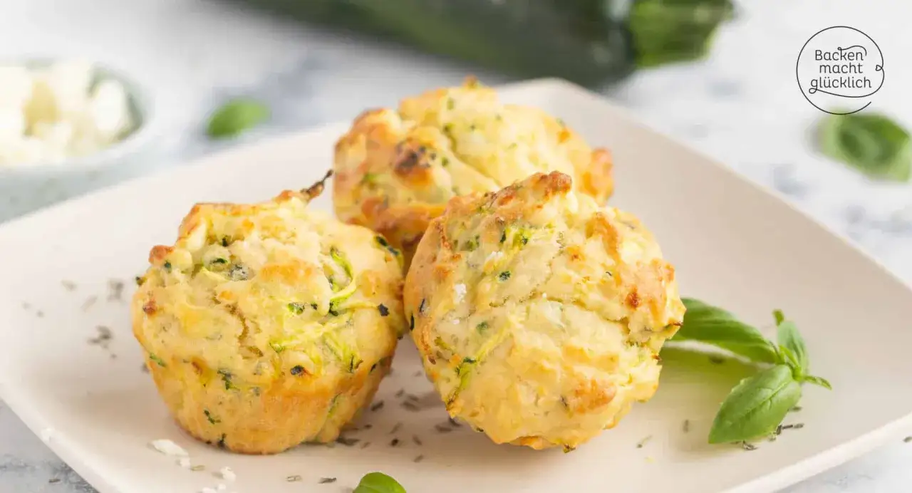 Drei goldbraune Zucchini-Muffins mit Kr&auml;utern und K&auml;se auf einem Teller, garniert mit Basilikum.